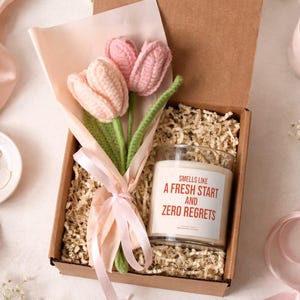 May include: A gift box containing a crochet tulip bouquet and a scented candle. The candle has a label that reads "Smells Like A Fresh Start And Zero Regrets". The box is filled with shredded paper and tied with a pink ribbon.