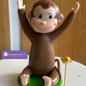 May include: A brown monkey figurine with arms raised, smiling, and seated on a green base. A small yellow bird on a stick is next to the monkey. The figurine is likely a cake topper or decorative item.