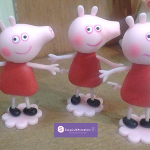 Puede incluir: Tres figuras de cerdos rosas con vestidos rojos y zapatos negros, de pie sobre bases en forma de flor rosa claro. Los cerdos tienen caras sonrientes y brazos extendidos. Probablemente sean juguetes o artículos de colección.