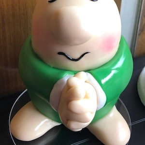 May include: A handcrafted porcelain figurine of a bald, smiling character with closed eyes and rosy cheeks. The figure wears a green tunic with white trim and clasped hands. It is displayed on a clear, round base.