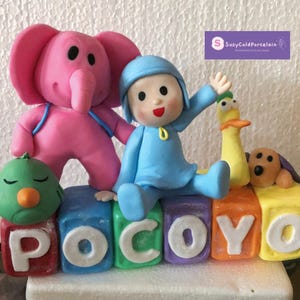 May include: A colorful cake topper featuring the cartoon character Pocoyo, an elephant, a duck, and a dog. The characters are arranged on blocks spelling "Pocoyo". The elephant is pink, Pocoyo is in blue, and the duck is yellow.