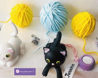 May include: A whimsical scene featuring cat figurines, yarn balls, and toy mice. The cats are white and black, with yellow eyes. The yarn balls are yellow and blue. A tag with the name "Juanita" is also present.