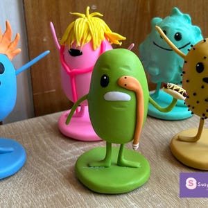 Puede incluir: Una colección de figuras de dibujos animados coloridas. Las figuras tienen forma de huevo con brazos y piernas, y cada una tiene un diseño único, incluyendo una figura azul con pelo naranja, una verde con una serpiente y una amarilla con un perrito caliente.