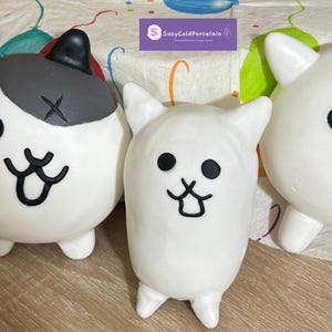 May include: White ceramic cat figurines with black facial features and small legs. One cat wears a black hat. A small penguin figurine is also present. The figurines are on a patterned surface.