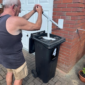 Peut inclure: Une personne utilise un ouvre-poubelle en métal blanc fixé à une poubelle noire. La poubelle porte le numéro 4. L'ouvre-poubelle est fixé sur un mur en briques rouges.