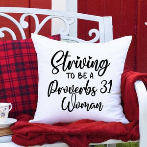 May include: White throw pillow with black text that reads "Striving to be a Proverbs 31 Woman". The pillow is on a white bench with a red and black plaid pillow and a red throw blanket.