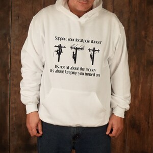 May include: White hooded sweatshirt with a black graphic of three linemen working on power lines. The text reads "Support your local pole dancer. It's not all about the money. It's about keeping you turned on."