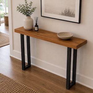 May include: A rectangular console table featuring a thick, natural-toned wooden top supported by two black metal U-shaped legs. The table surface holds a white ceramic vase with green foliage, a small dark glass diffuser, and a shallow beige bowl.