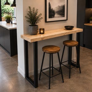 May include: A rectangular bar table with a light wood top and black metal legs, accompanied by two round wooden bar stools with black metal frames. A potted green plant and a small glass candle holder sit on the table surface.