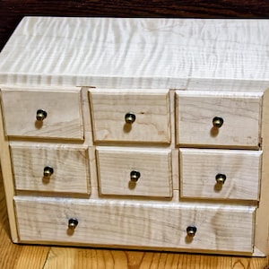 May include: A small, light-colored wooden chest of drawers with nine drawers. Each drawer has a small, round, brass-colored knob. The wood grain is visible on the chest and drawers. The chest is rectangular with rounded edges.
