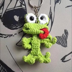 Könnte beinhalten: Ein handgefertigter grüner Häkelfrosch-Schlüsselanhänger mit weißen Augen und einer roten Zunge. Der Frosch hat einen Schlüsselring an seinem Kopf befestigt. Der Hintergrund ist eine graue Oberfläche mit einem schwarz-weißen Design.