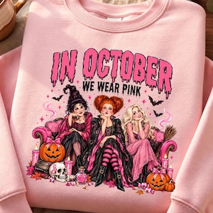 Op de afbeelding: Een lichtroze sweater met ronde hals en een grafische print van drie heksen die op een roze bank zitten. De tekst "IN OCTOBER WE WEAR PINK" staat in een druipend lettertype. Het ontwerp bevat pompoenen, een schedel met een roze lint en vleermuizen.