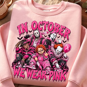 Op de afbeelding: Een lichtroze sweater met ronde hals en een grafische print van vijf horrorfilmfiguren met roze accenten en lintjes voor borstkankerbewustzijn. De tekst luidt IN OCTOBER WE WEAR PINK in een druipend, vet, felroze lettertype.