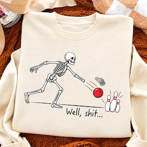 Op de afbeelding: Een crèmekleurig sweatshirt met ronde hals en een afbeelding van een skelet dat aan het bowlen is. Het skelet heeft een rode bowlingbal naar drie witte kegels gegooid. De hand van het skelet is losgekomen van de arm. De tekst luidt, Well, shit...