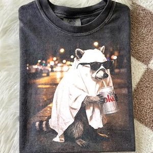 Op de afbeelding: Een antracietgrijs T-shirt met ronde hals en een grafische print van een wasbeer die een wit laken als spookkostuum en een zwarte zonnebril draagt, terwijl het dier een zilverkleurig blikje Diet Coke vasthoudt tegen een stadsachtergrond.