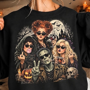 Op de afbeelding: Een zwart sweatshirt met ronde hals en een grafische print van drie vrouwen met rood, blond en donker haar die een zonnebril dragen, naast een groen monsterfiguur. Het ontwerp bevat een volle maan, een kasteel, vleermuizen, een pompoen en een schedel.