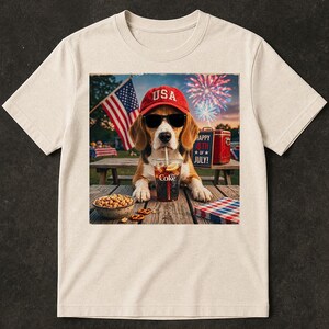 Op de afbeelding: Een cr&egrave;mekleurig t-shirt met een afbeelding van een beagle met een rode USA-pet en zwarte zonnebril, drinkend uit een Coke-beker. De sc&egrave;ne bevat een schaaltje pinda's, een rode koelbox met Happy 4th of July, een Amerikaanse vlag en vuurwerk.