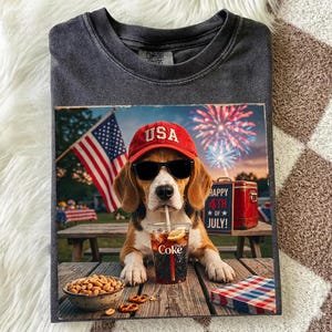 Op de afbeelding: Een antracietgrijs T-shirt met ronde hals en een afbeelding van een beagle met een rode USA-honkbalpet en een zwarte zonnebril. De hond houdt een rietje in een glas Diet Coke. Op de achtergrond zijn een Amerikaanse vlag, vuurwerk en een bord met HAPPY 4TH OF JULY te zien.