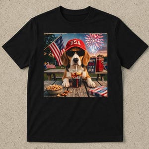 Op de afbeelding: Een zwart t-shirt met een afbeelding van een beagle met een rode USA-pet en zwarte zonnebril, drinkend uit een Coke-beker met een rietje. De afbeelding bevat een kom pretzels, een Amerikaanse vlag, vuurwerk en de tekst Happy 4th of July.