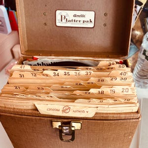 May include: Vintage brown Amfile Platter-pak record storage case, open to reveal numbered file dividers. The case has a metal clasp and a label that reads "Amfile Platter-pak". The dividers are tan with black numbers.