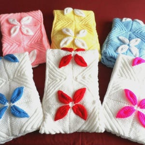 May include: A collection of six knitted decorative pillows in various colors, including pink, yellow, and blue. Each pillow features a central floral design in contrasting colors, such as white, red, and pink. The pillows are arranged on a red surface.