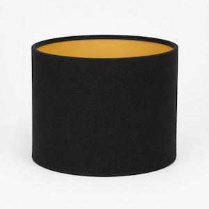 May include: A cylindrical drum lampshade featuring a solid black fabric exterior with a fine woven texture. The interior lining of the lampshade is a contrasting solid mustard yellow color, visible through the open top and bottom circular openings.