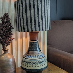 May include: A ceramic table lamp with a striped lampshade. The base is a mix of blue, brown, and beige with a decorative pattern. The lamp is on a wooden table next to a vase and a copper dish.