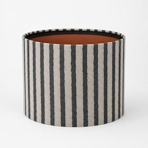 May include: A cylindrical lampshade featuring a vertical striped design in black and beige. The interior is a warm brown, and the top edge is finished with a black band. This lampshade is suitable for home lighting.