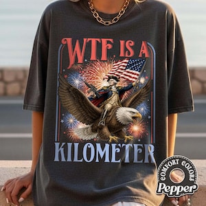 May include: Dark gray t-shirt with a patriotic design. The graphic features a man riding an eagle, holding a flag, with fireworks and the text "WTF IS A KILOMETER". The shirt has a Comfort Colors label.