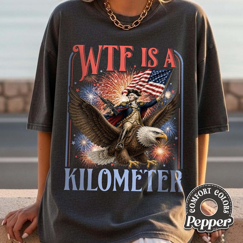 May include: Dark gray t-shirt with a patriotic design. The graphic features a man riding an eagle, holding a flag, with fireworks and the text "WTF IS A KILOMETER". The shirt has a Comfort Colors label.