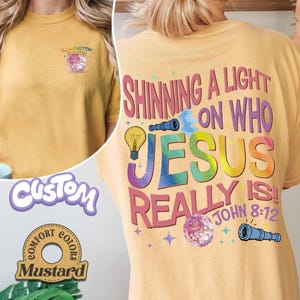 May include: Mustard-colored t-shirt with the text "SHINNING A LIGHT ON WHO JESUS REALLY IS! JOHN 8:12" in a rainbow design. The front of the shirt has the text "ILLUMINATION SESSION" and a graphic. The shirt is a Comfort Colors brand.