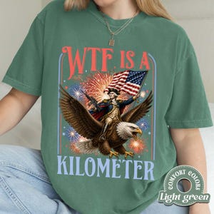 May include: Light green t-shirt with a patriotic design. The graphic features a man riding an eagle, holding a rifle and an American flag, with the text "WTF IS A KILOMETER" in red and blue. Fireworks are in the background.
