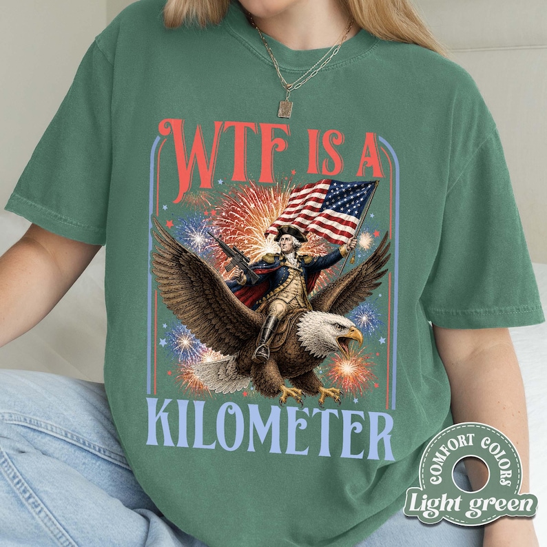 May include: Light green t-shirt with a patriotic design. The graphic features a man riding an eagle, holding a rifle and an American flag, with the text "WTF IS A KILOMETER" in red and blue. Fireworks are in the background.