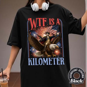 May include: Black t-shirt with a graphic featuring an eagle carrying a figure holding a flag and a rifle, with fireworks in the background. The text reads "WTF IS A KILOMETER". The shirt has a Comfort Colors label.