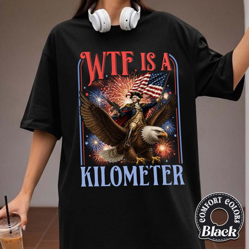 May include: Black t-shirt with a graphic featuring an eagle carrying a figure holding a flag and a rifle, with fireworks in the background. The text reads "WTF IS A KILOMETER". The shirt has a Comfort Colors label.