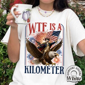May include: White t-shirt with a graphic featuring a man riding an eagle, holding a flag and a rifle, with fireworks and the text "WTF IS A KILOMETER". The shirt has a "Comfort Colors" label.