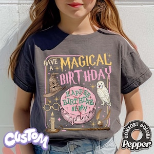 May include: Dark gray t-shirt with a graphic design that reads "Have a Magical Birthday" in yellow and pink. The design includes a sorting hat, glasses, a cake, an owl, a wand, and a golden snitch. The shirt has the word "Custom" on the bottom left.