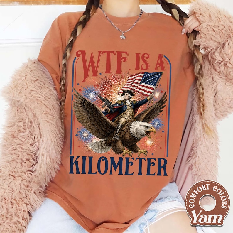 May include: Burnt orange t-shirt with a graphic of a man riding an eagle, holding a flag and a rifle, with fireworks in the background. Text reads "WTF IS A KILOMETER". The shirt has a Comfort Colors tag.