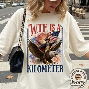 May include: Ivory-colored t-shirt with a graphic featuring an eagle carrying a figure with an American flag. The text reads "WTF IS A KILOMETER". The design includes fireworks and patriotic colors.