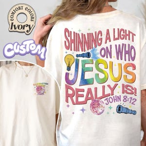 May include: Ivory-colored t-shirt with the text "Shining a Light on Who Jesus Really Is!" in a colorful, rainbow design. The shirt also features a small graphic on the front. The shirt is a Comfort Colors brand.
