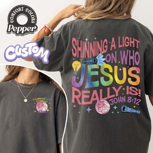 May include: Dark gray Comfort Colors t-shirt with the text "Shining a Light on Who Jesus Really Is!" in a colorful design. The front of the shirt has the text "Illumination Station" and a graphic. The shirt also has the text "John 8:12".