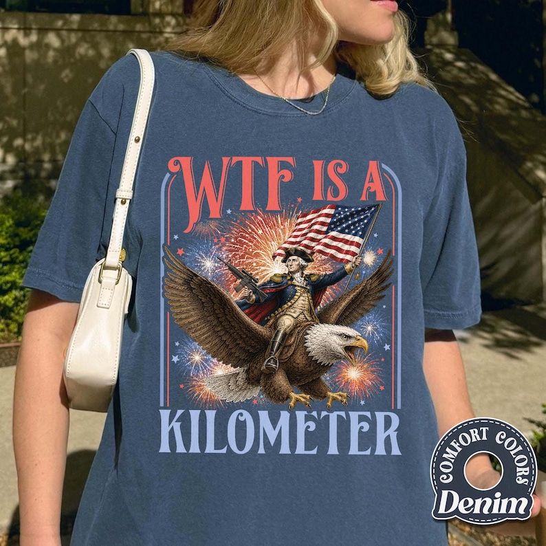 May include: Navy blue t-shirt with a graphic featuring an eagle carrying a man holding a flag, with fireworks in the background. The text reads "WTF IS A KILOMETER". The shirt is made of Comfort Colors denim.