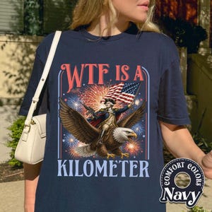 May include: Navy blue t-shirt with a graphic featuring an eagle carrying a figure holding a flag, with fireworks in the background. The text "WTF IS A KILOMETER" is displayed above and below the image. The shirt has a "Comfort Colors" logo.