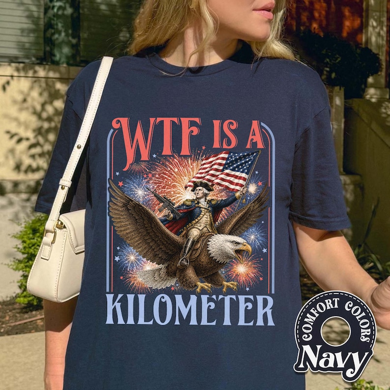 May include: Navy blue t-shirt with a graphic featuring an eagle carrying a figure holding a flag, with fireworks in the background. The text "WTF IS A KILOMETER" is displayed above and below the image. The shirt has a "Comfort Colors" logo.