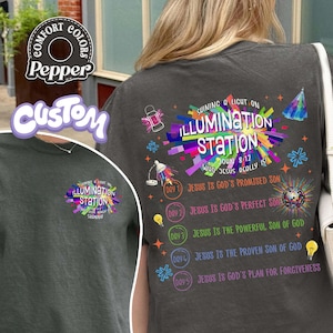 K&ouml;nnte beinhalten: Ein dunkelgraues T-Shirt mit einer bunten Grafik auf der R&uuml;ckseite und einer kleineren Version auf der Vorderseite. Das Design enth&auml;lt den Text Shining a Light on Illumination Station, John 8:12 und eine Liste von f&uuml;nf Tagesthemen mit Lichtsymbolen.