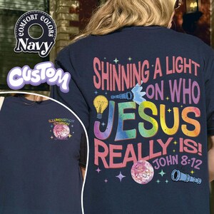 May include: Navy blue t-shirt with the text "Shining a Light on Who Jesus Really Is! John 8:12" in colorful letters. The shirt also features a lightbulb, telescope, and disco ball graphic. A smaller image on the front says "Illumination Station".