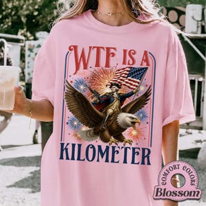 May include: Pink t-shirt with a graphic of a man riding an eagle, holding a flag, and the text "WTF IS A KILOMETER." Fireworks and patriotic colors are in the background. The shirt is a Comfort Colors brand.