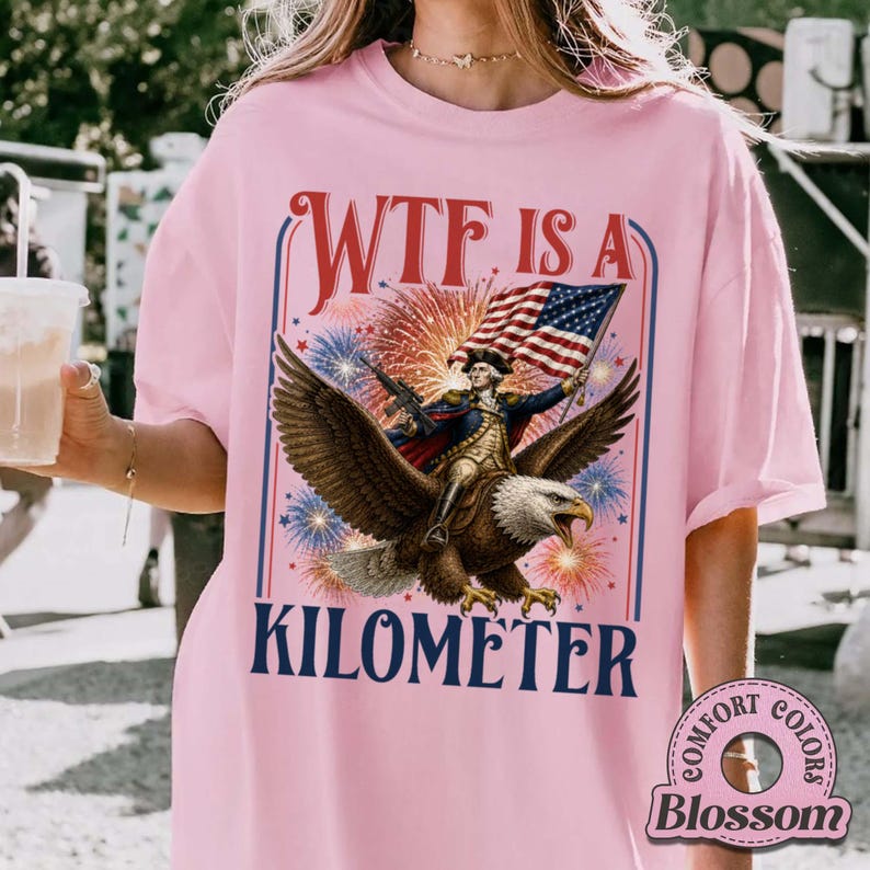 May include: Pink t-shirt with a graphic of a man riding an eagle, holding a flag, and the text "WTF IS A KILOMETER." Fireworks and patriotic colors are in the background. The shirt is a Comfort Colors brand.