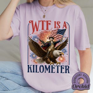 May include: A light purple t-shirt with a graphic featuring a man riding an eagle, holding an American flag, and surrounded by fireworks. The text "WTF IS A KILOMETER" is displayed in a bold font.