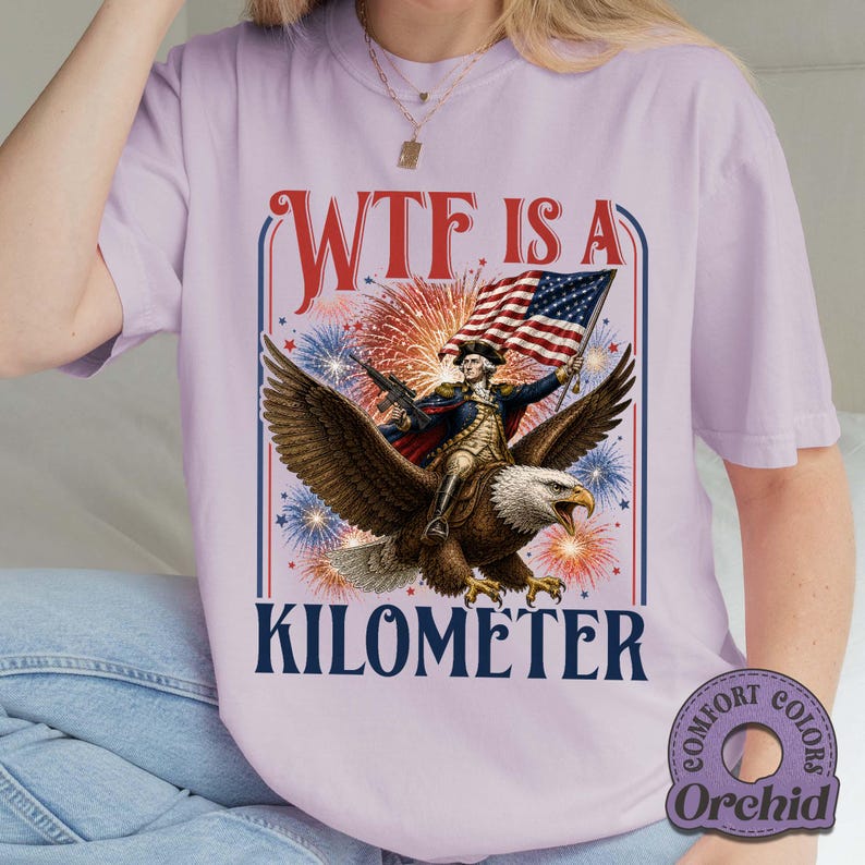 May include: A light purple t-shirt with a graphic featuring a man riding an eagle, holding an American flag, and surrounded by fireworks. The text "WTF IS A KILOMETER" is displayed in a bold font.